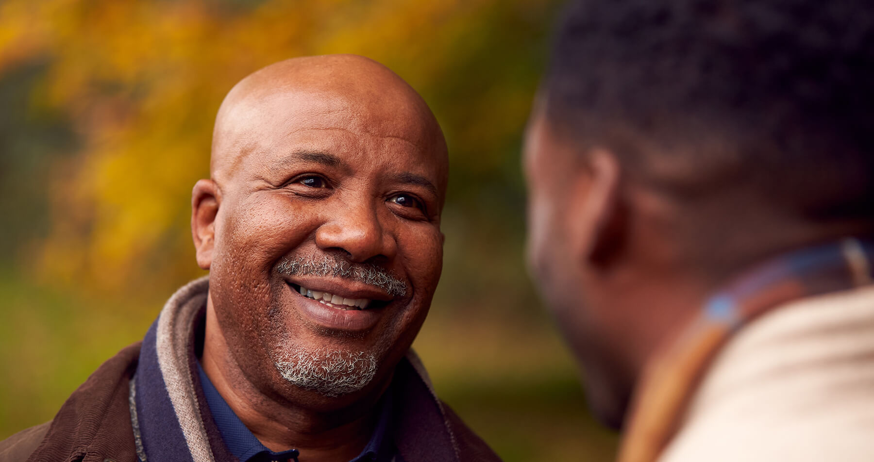 A Black father talking with his adult son, making direct eye contact, a caring smile engaging laugh lines. His son’s partial profile hints at a smile