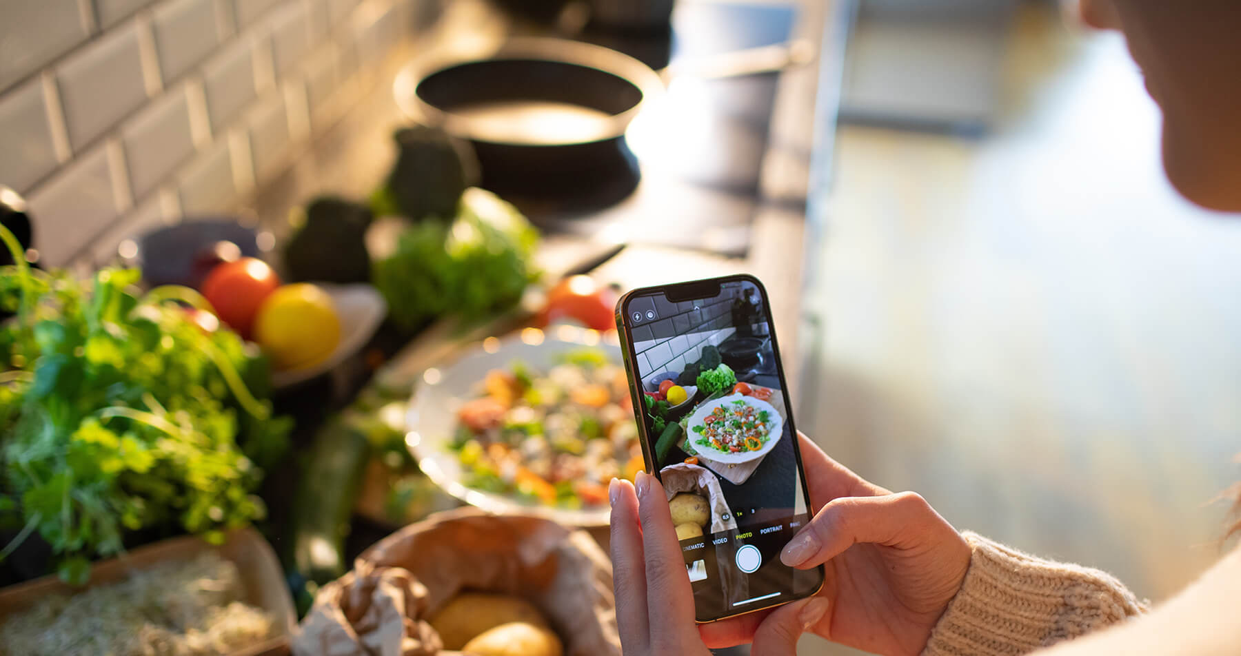 Photo of a woman holding a cell phone taking pictures of food on her kitchen counter in various states of preparation