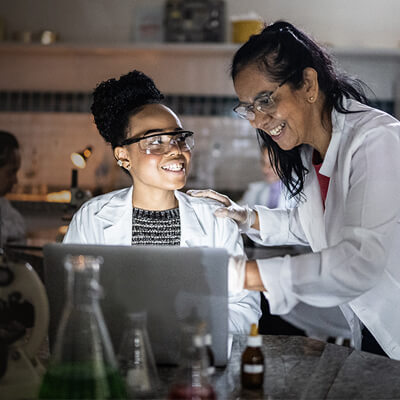 In a lab, a woman of East Asian decent smiles, pointing at something on the computer in front of a seated Black woman, who smiles back at her mentor