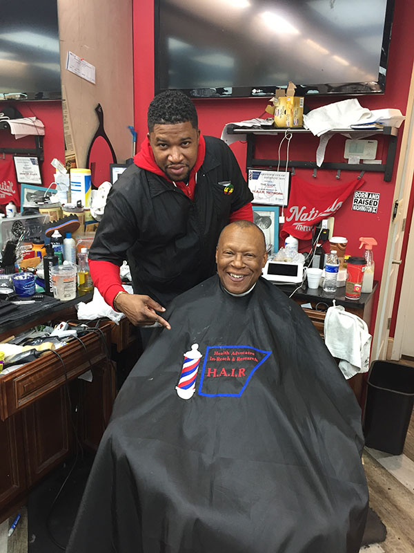 Dr. Thomas sits in a barbershop chair. Fred Spry stands behind him pointing to Dr. Thomas’ HAIR-branded cape.  