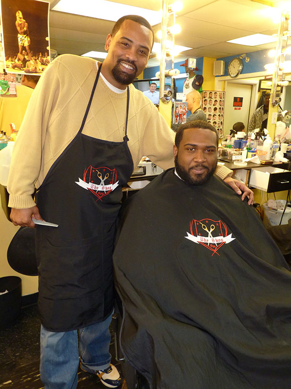 Michael Brown holds a comb and has his arm around a customer who is sitting in a barber chair. 