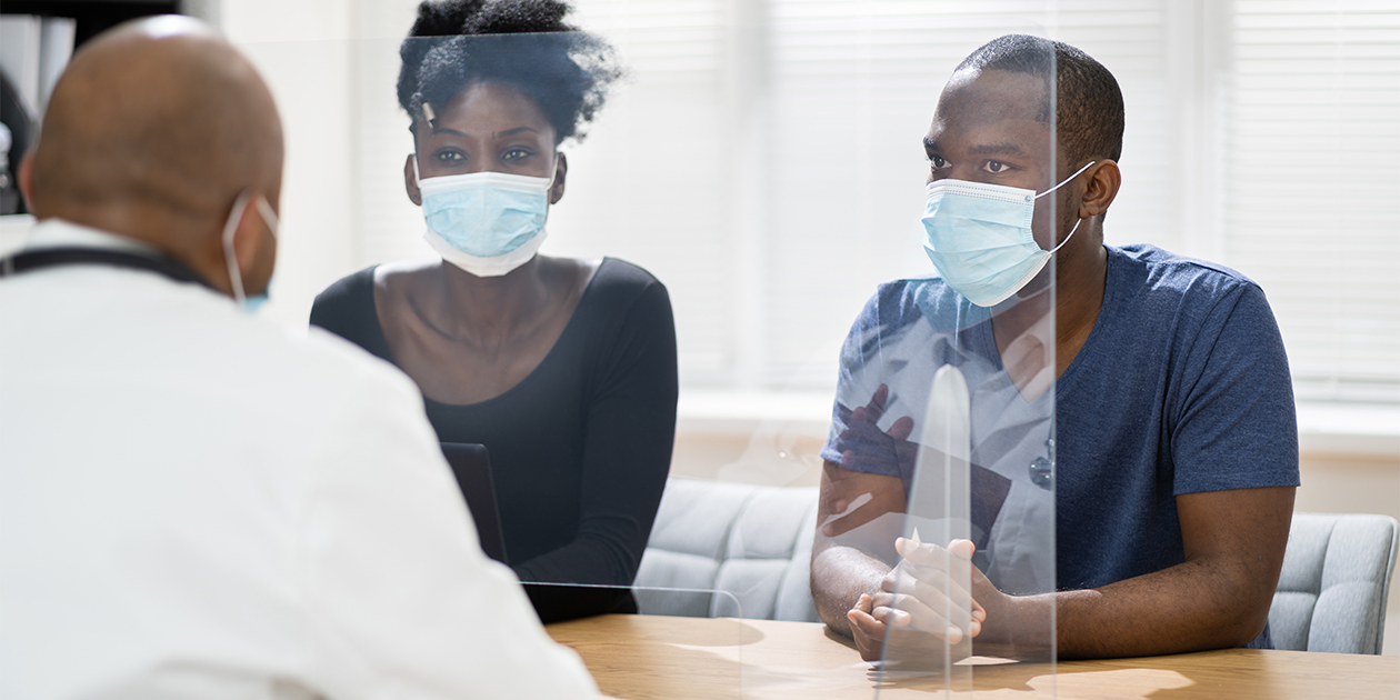 African American male doctor wearing mask and a white coat and sitting behind a clear, plexiglass divider while talking to two African American patients (one male and one female), both wearing blue medical facemasks.