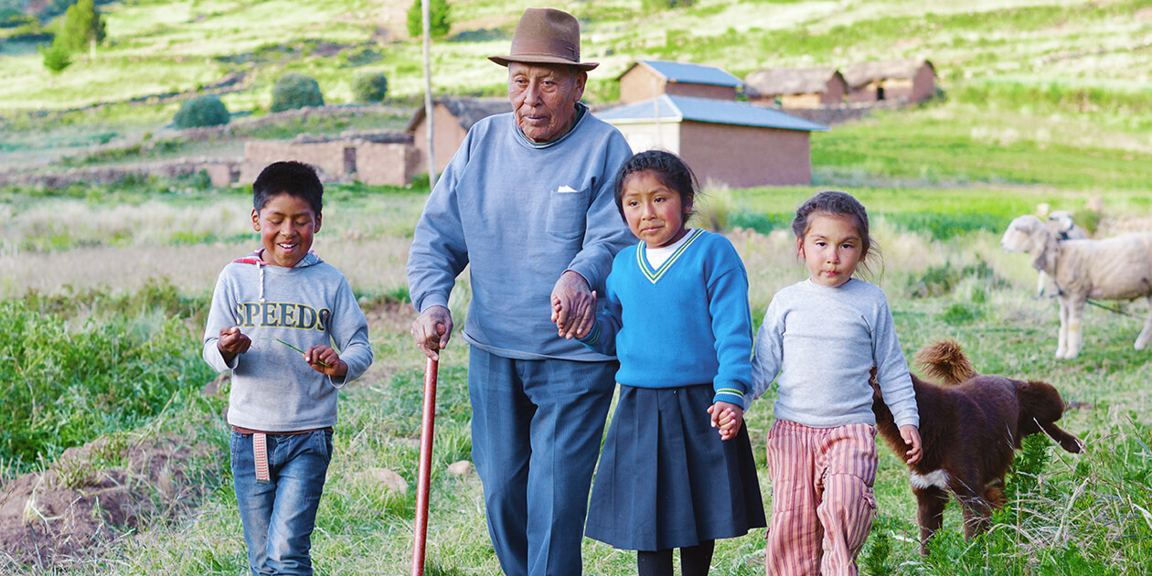 A Native American man using a walking stick walks with his 3 grandchildren in the countryside. Farm animals and low buildings are in the background