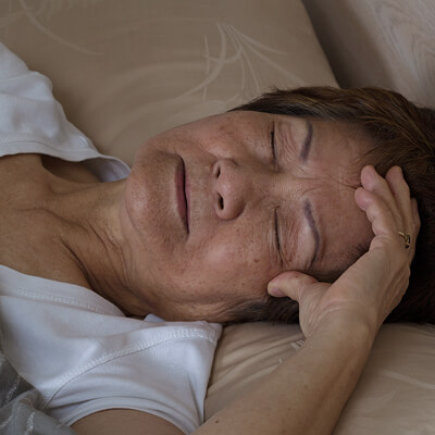 Photo showing the head and shoulders of an older woman of Asian descent sleeping. She has 1 hand on her forehead, her brow is slightly furrowed