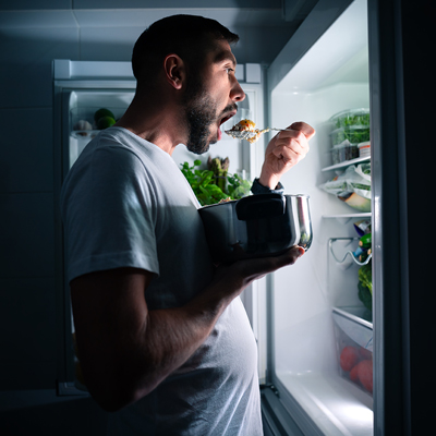 Photo of a man standing in a darkened kitchen in front of an open refrigerator holding a pot of food with a spoon of food to his mouth