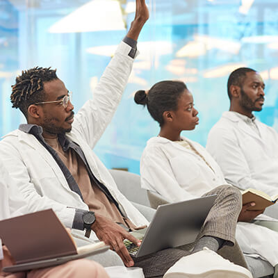 Medical students, 2 Black men, 1 with his hand raised to speak, and a Black woman in a classroom.