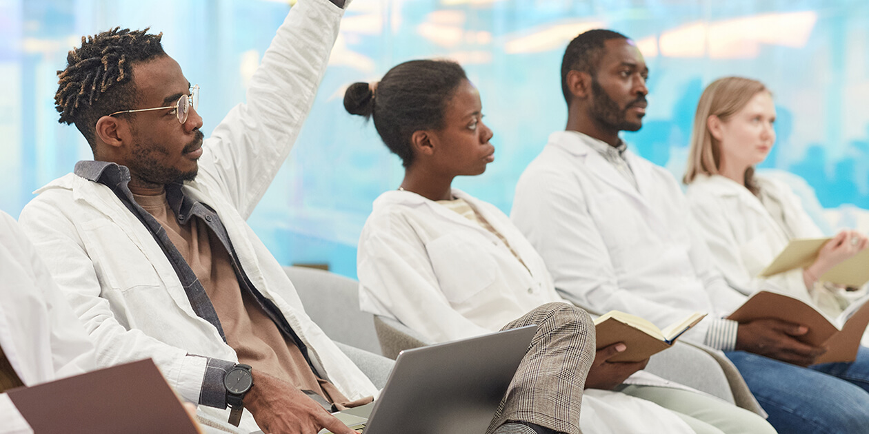Medical students, 2 Black men, 1 with his hand raised to speak, a Black woman and a White woman in a classroom